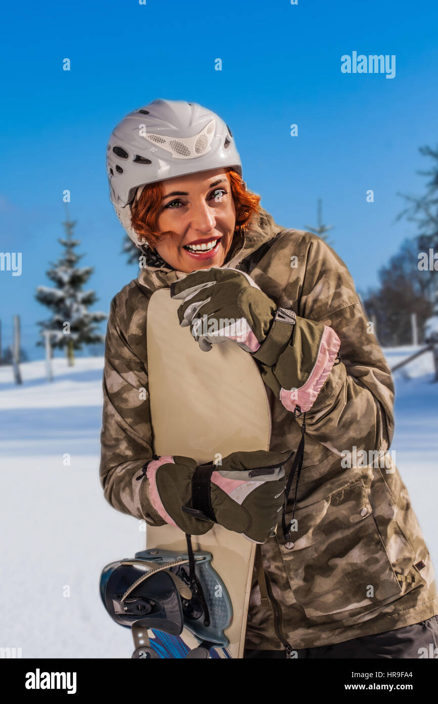 La parte superiore del corpo in aspirazione clima soleggiato di un redheaded, ridendo donna con casco, in piedi sulle piste con lo snowboard in mano. Foto Stock