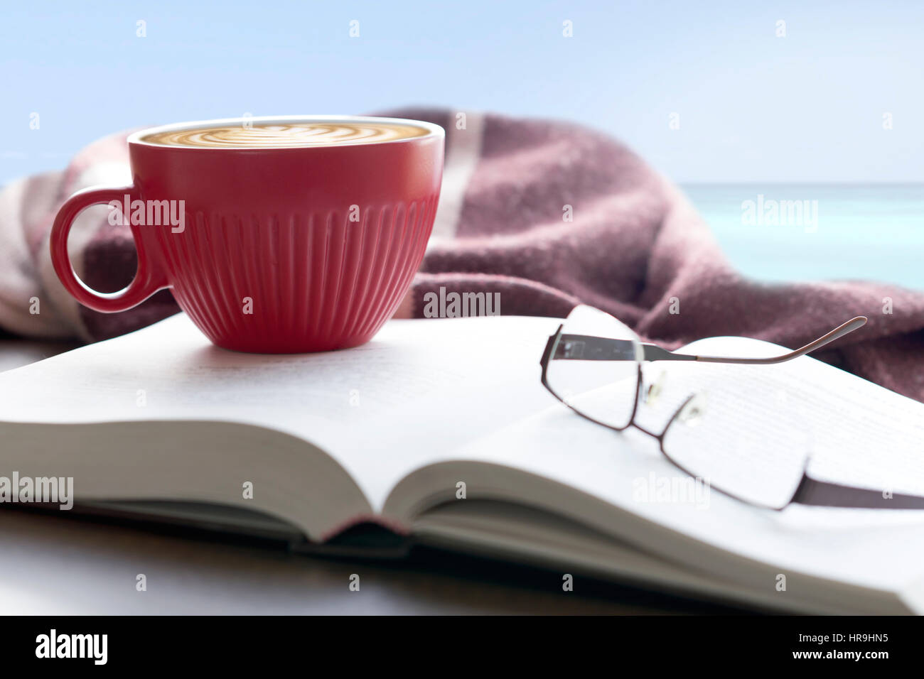 Pausa caffè al mare, rosso tazza di caffè sul libro con gli occhiali da lettura e un maglione in riva al mare Foto Stock