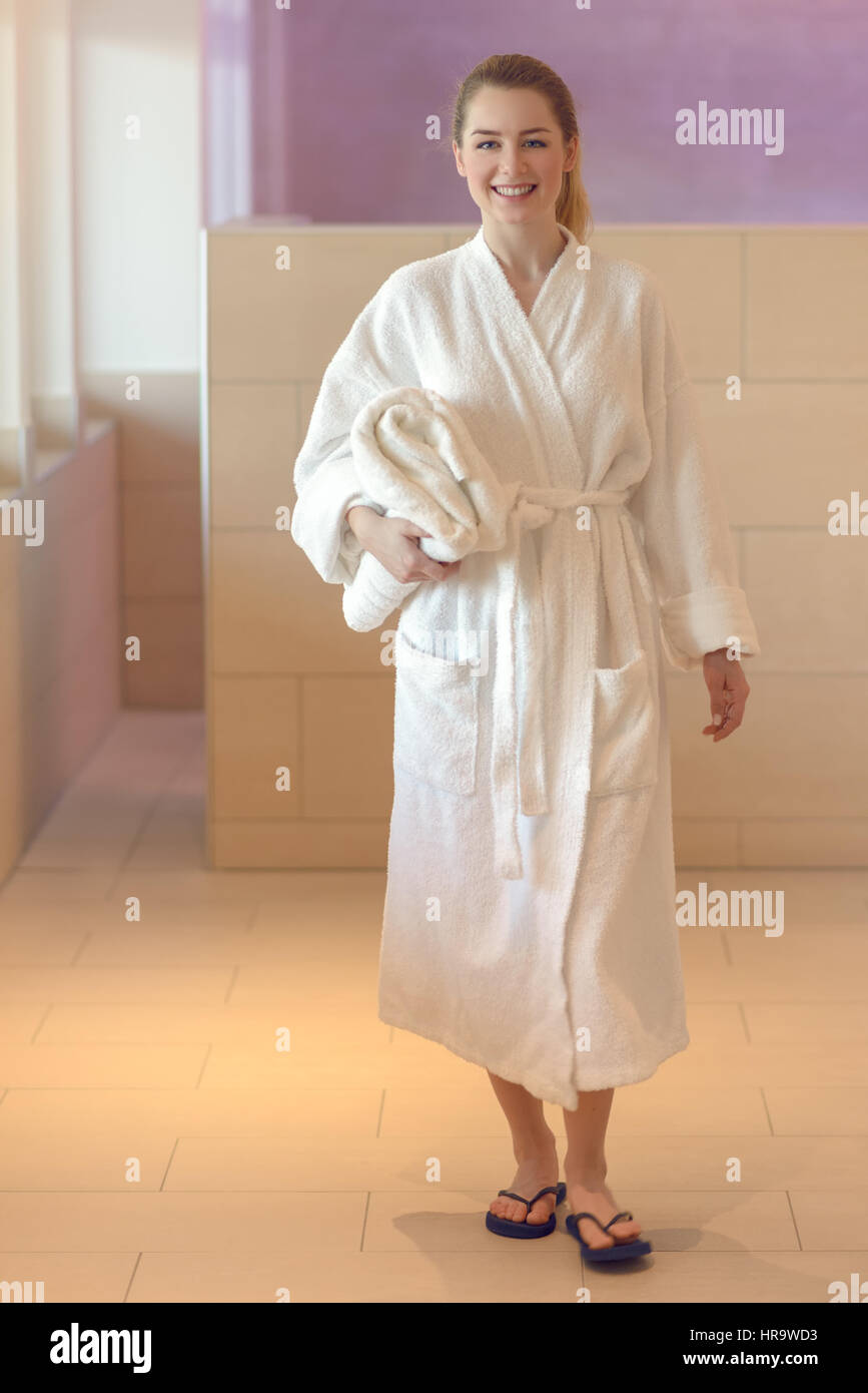 Piuttosto giovane donna pronta a prendere una doccia o un bagno in piedi in un bianco accappatoio in spugna tenendo una salvietta ripiegata sotto il suo braccio come sorride a camma Foto Stock
