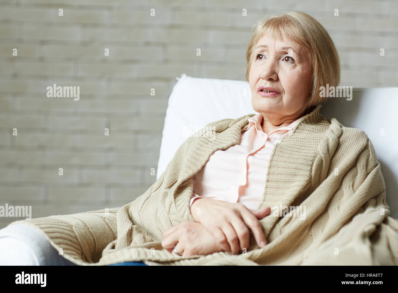 Vita-up verticale di pensieroso bionda donna senior di rosa pallido camicetta seduti sul comodo divano e coperto le spalle con brown plaid Foto Stock