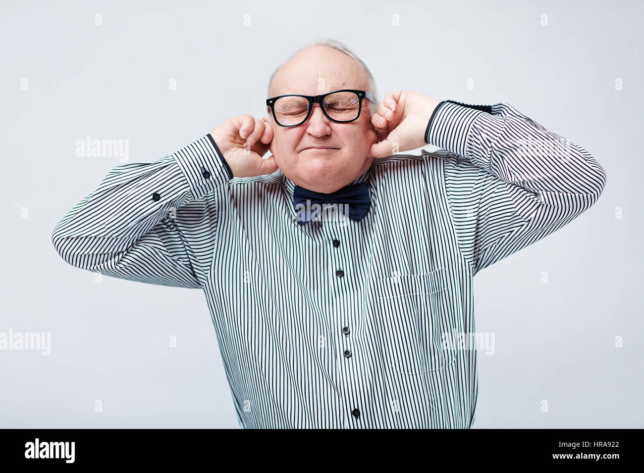 Vita-up verticale di elegante uomo anziano in camicia a righe con il filtro bow tie collegando le sue orecchie con le dita e spremendo i suoi occhi strettamente Foto Stock
