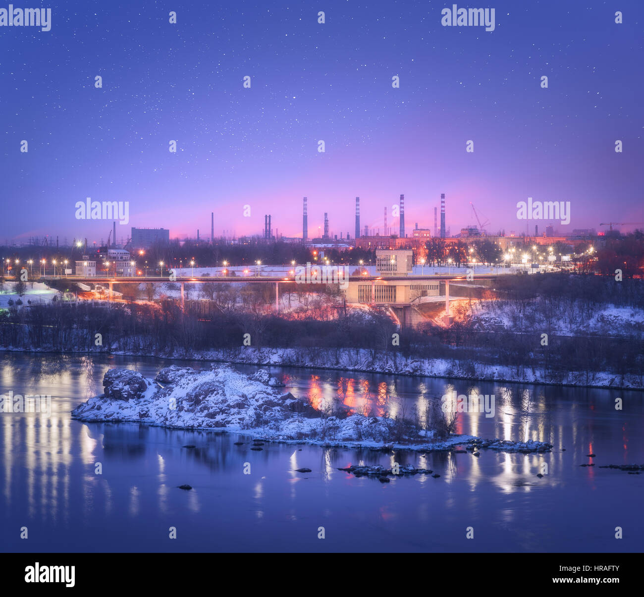 Notte cityscape con colorate cielo viola con stelle, rocce, fiume, alberi, edifici, illuminazione della città e fabbrica di acciaio con fumaiolo in inverno Foto Stock