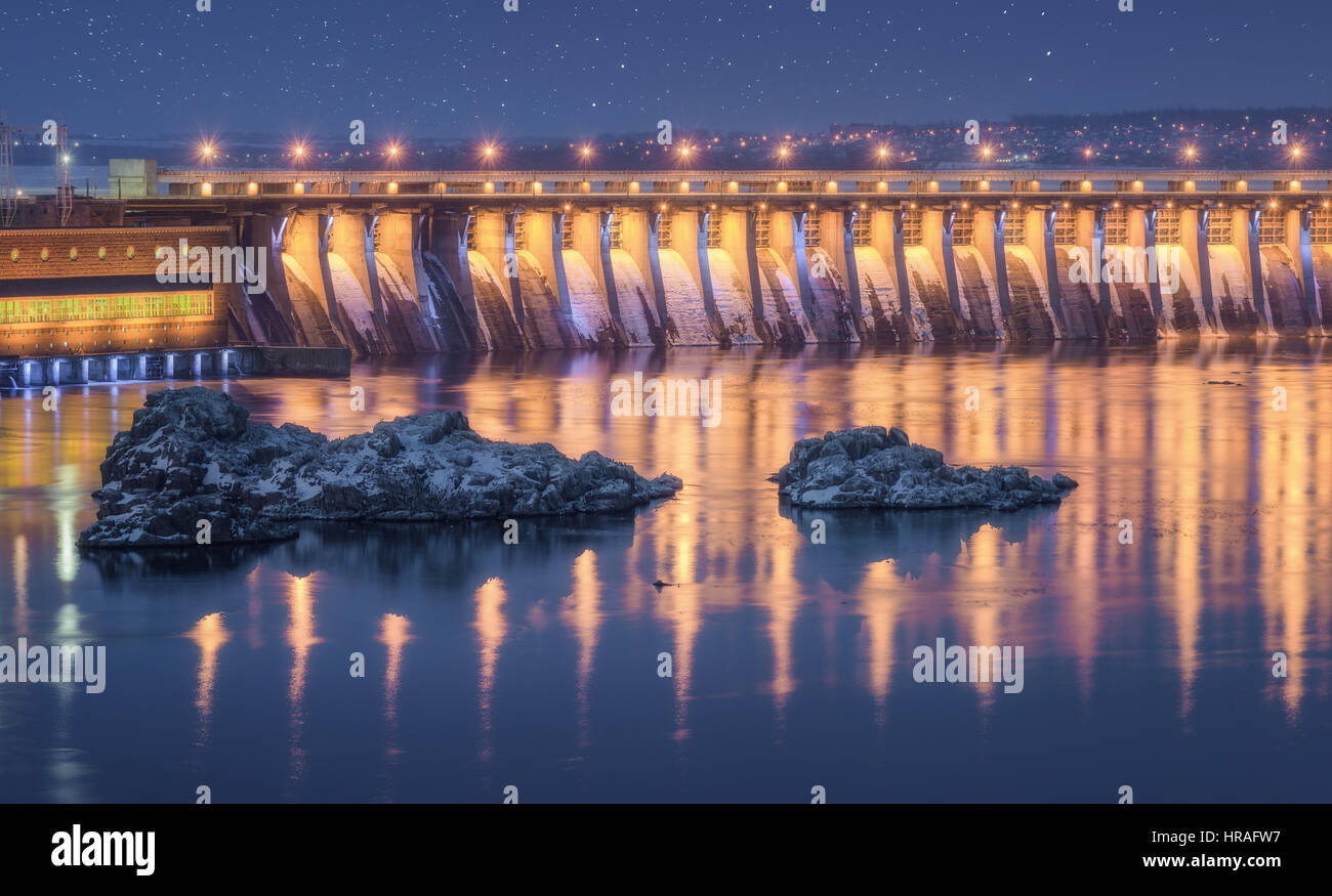 Diga. Bella notte paesaggio industriale con la diga idroelettrica, il ponte sul fiume, illuminazione della città riflessa nell'acqua, rocce Foto Stock