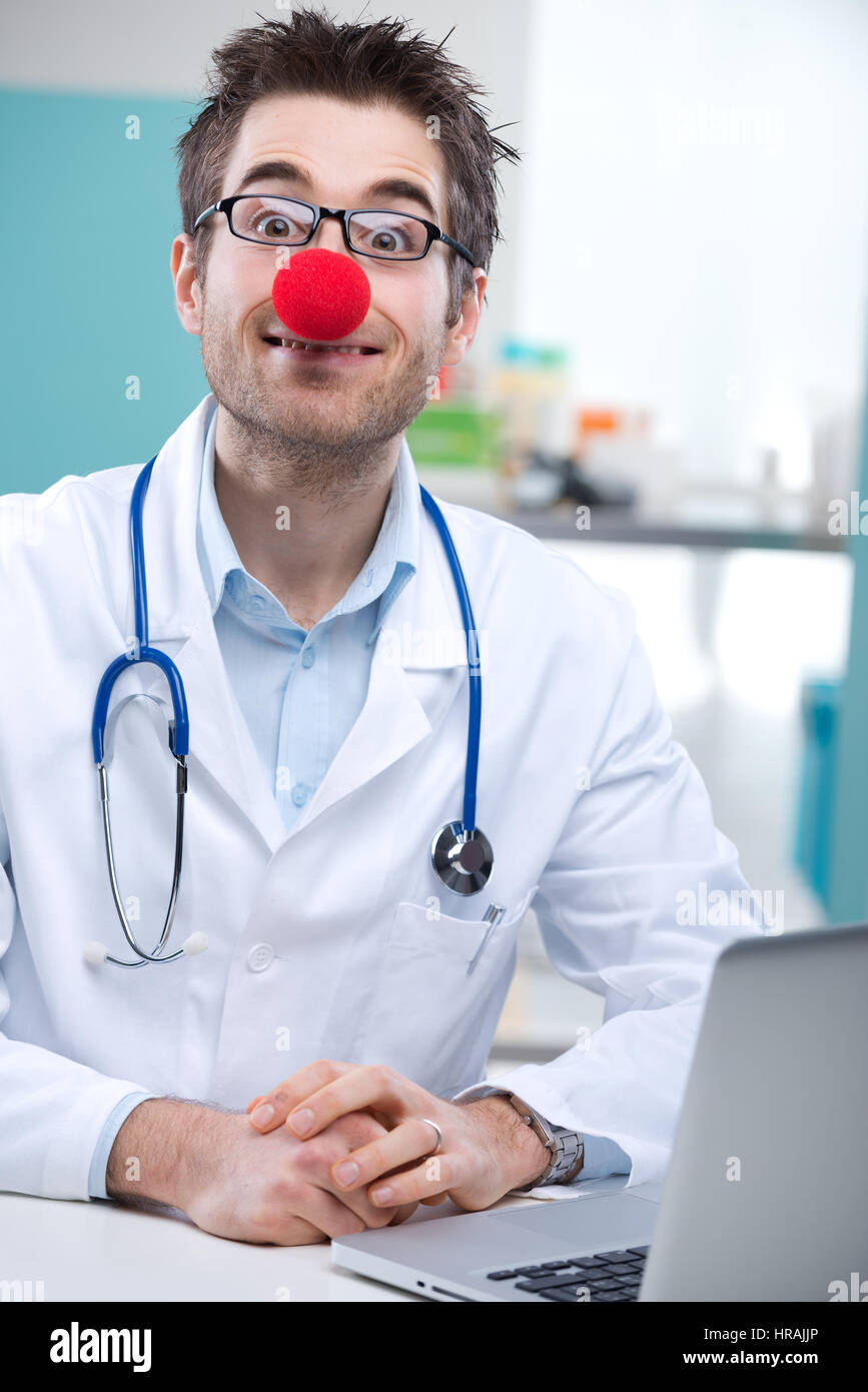 Funny giovane medico con un clown naso lavorando alla sua scrivania Foto Stock
