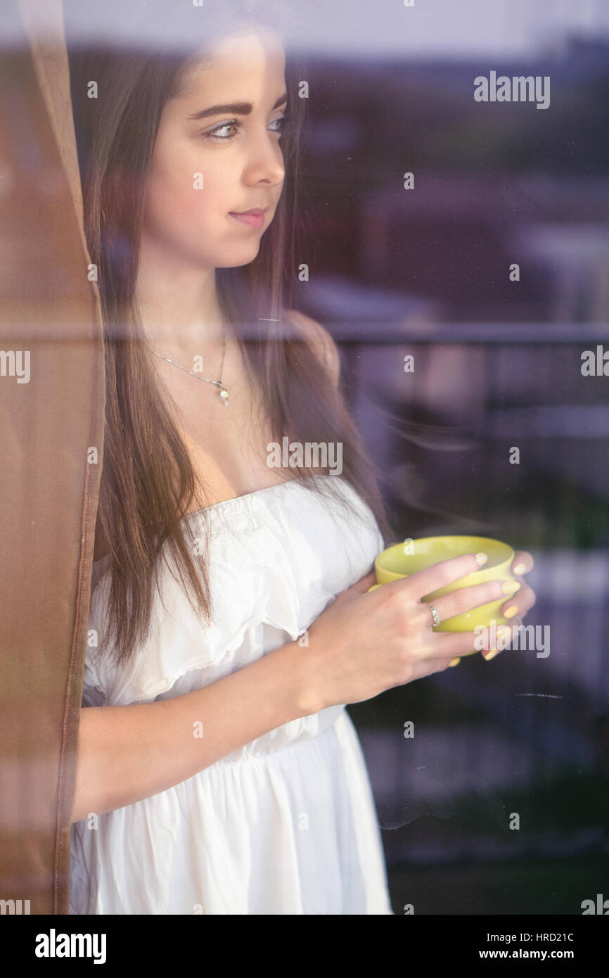 Bellissima giovane donna di bere il suo caffè fumante davanti alla finestra e guardando il ciity / gli effetti di colore e bilanciamento del bianco personalizzato viene aggiunto t Foto Stock