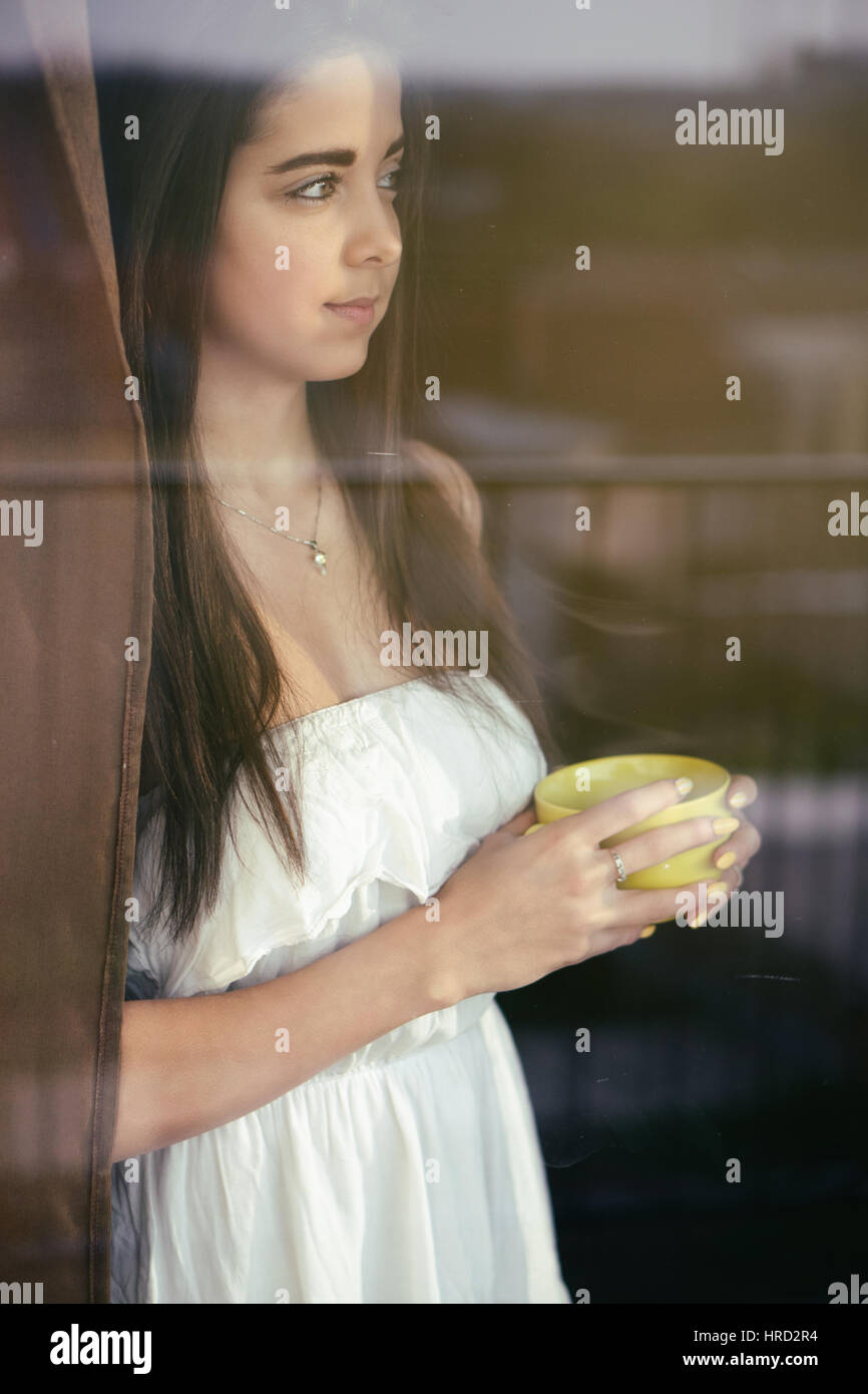 Bellissima giovane donna di bere il suo caffè fumante davanti alla finestra e guardando il ciity / gli effetti di colore e bilanciamento del bianco personalizzato viene aggiunto t Foto Stock