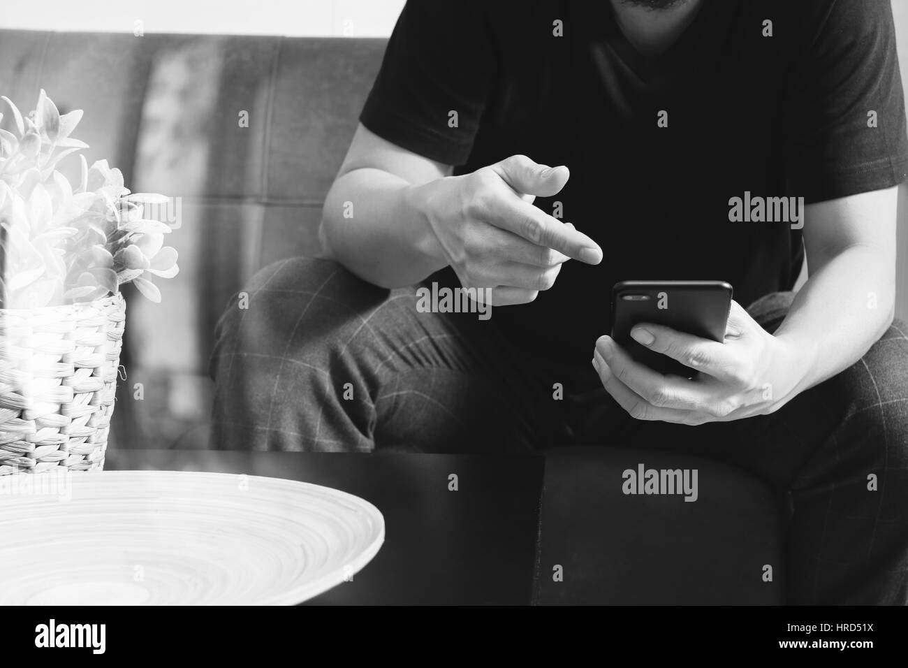 Designer man mano utilizzando smart phon per i pagamenti mobili shopping online,omni canale,seduto sul divano nel soggiorno,vaso rattan con impianti e t in legno Foto Stock