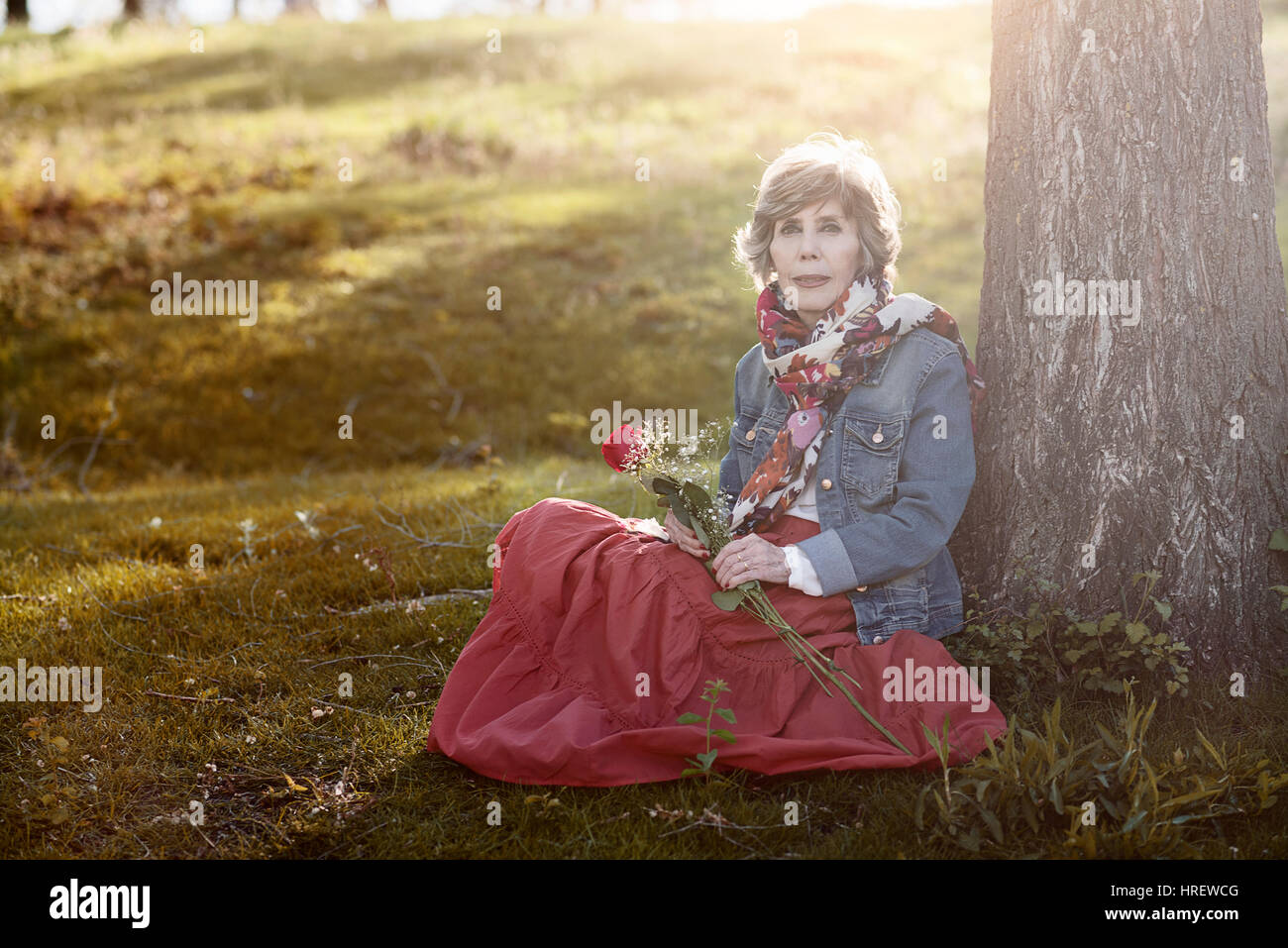 Felice senior donna seduta sull'erba. Senior donna con una rosa in mano Foto Stock