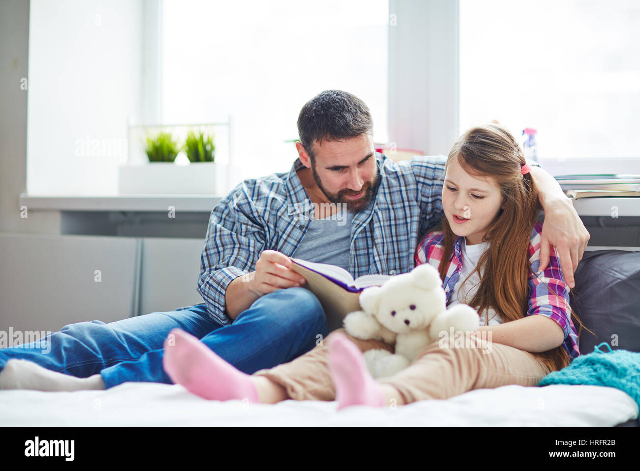 Ritratto a figura intera di pretty famiglia: bambina holding Teddy bear in mani durante la lettura del racconto eccitante di suoi barbuti di mezza età padre Foto Stock