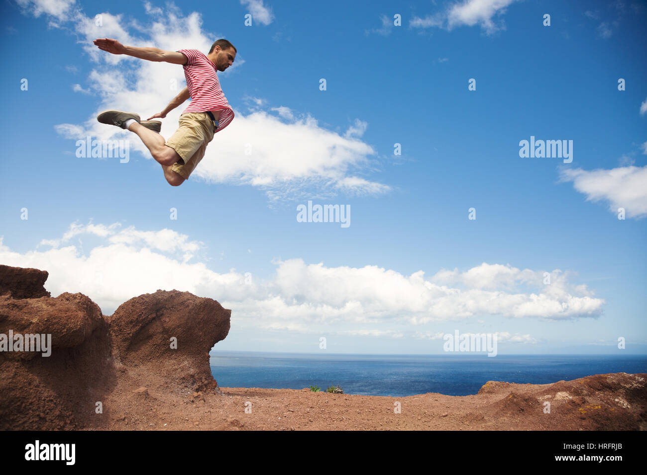 Giovane intrepido escursionista salta fuori la roccia con le gambe piegate su sfondo oceano, Tenerife Foto Stock