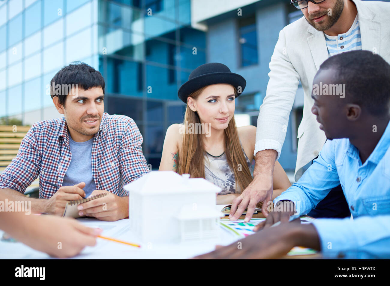 Multi-etnico gruppo di giovani architetti che lavorano insieme al tavolo esterno con modello di casa su di esso, discutendo di schema di colori per progetto di alloggiamento, collabor Foto Stock