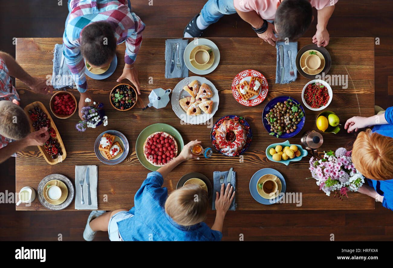 Famiglia avente pasto pomeridiano, seduti al grande tavolo in legno con gustosi appena sfornato dolci, giardino di bacche e frutta e tazze di tè Foto Stock
