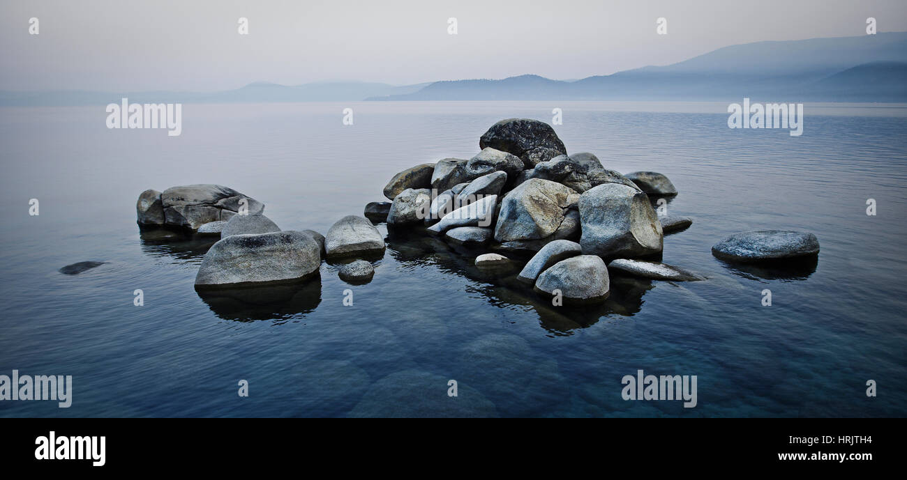 Un mucchio di pietre che si eleva al di sopra del livello di acqua in un lago in una nebbiosa paesaggio. Foto Stock