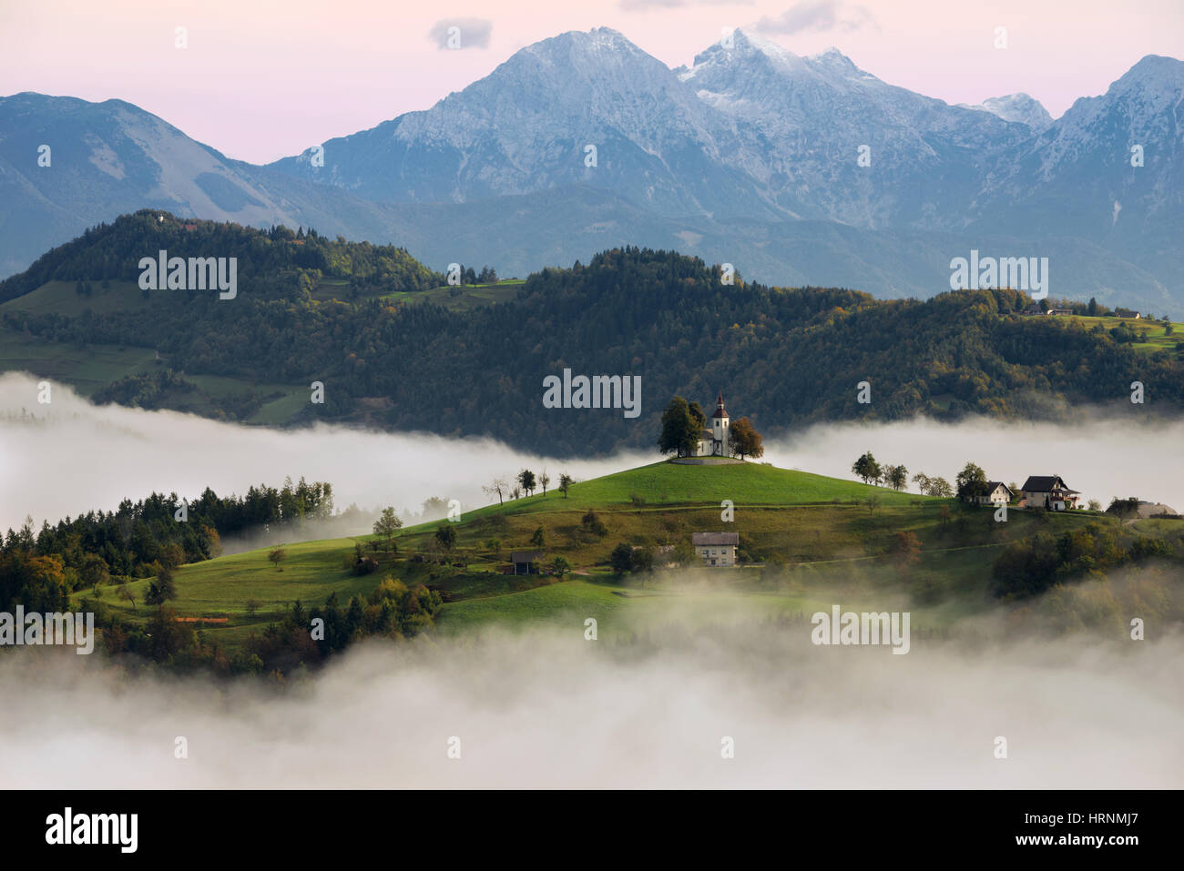 La chiesa di San Tommaso (Sveti Tomaz) sulla cima di una collina nella campagna slovena con la nebbia con sullo sfondo la neve sulle montagne in Slovenia. Foto Stock