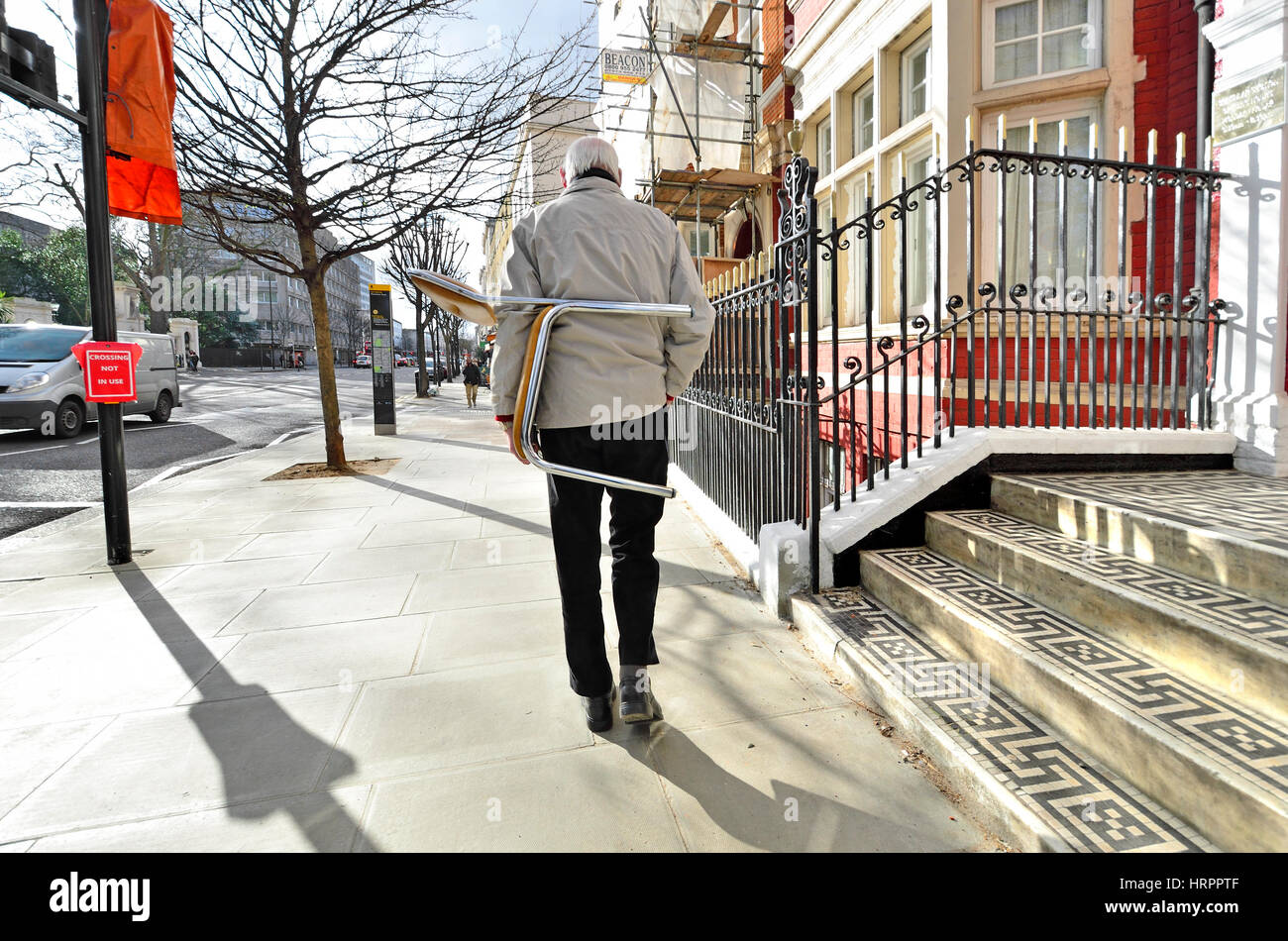 Londra, Inghilterra, Regno Unito. Kensington - uomo che porta una sedia in fondo alla strada Foto Stock