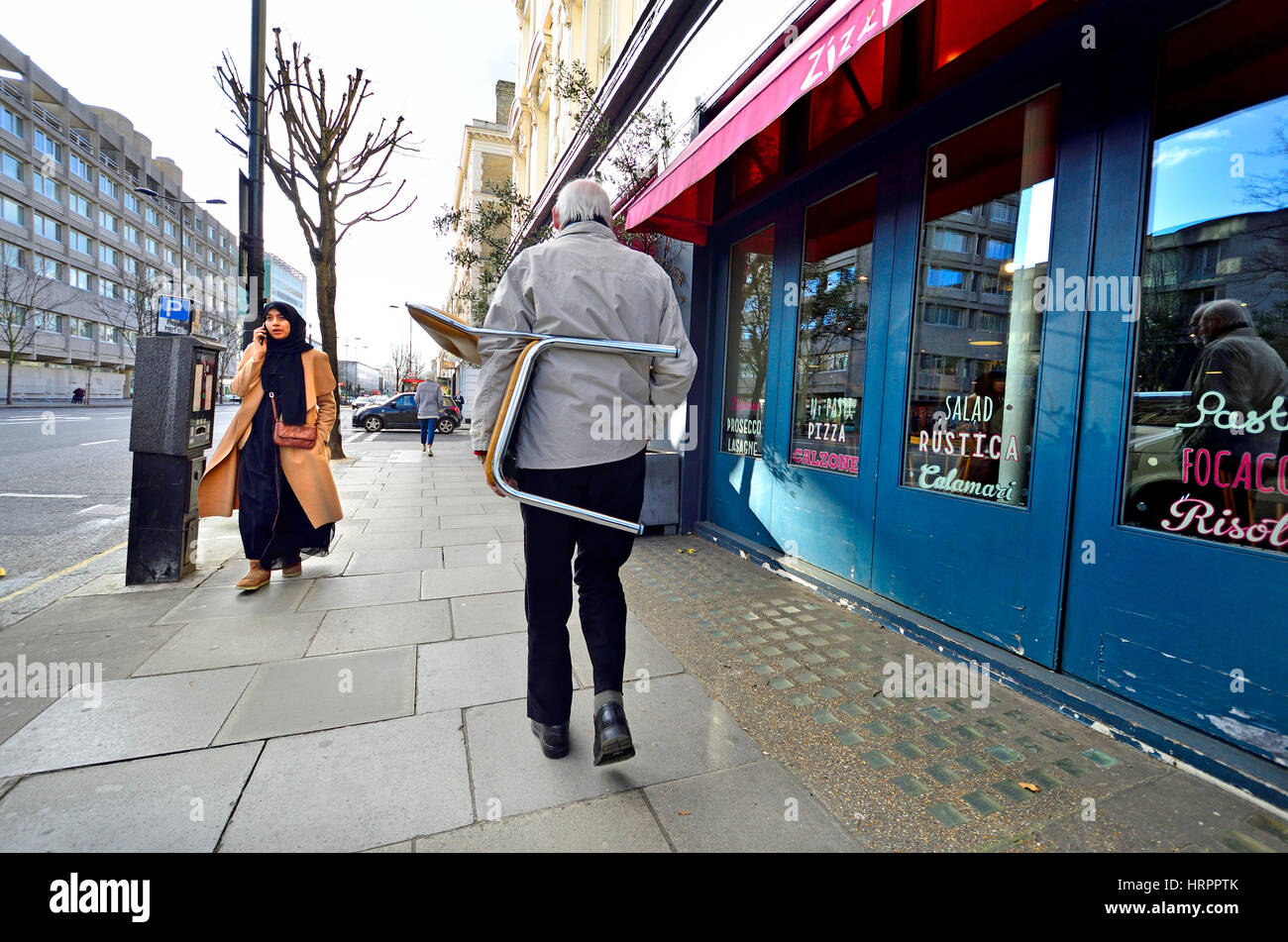 Londra, Inghilterra, Regno Unito. Kensington - uomo che porta una sedia in fondo alla strada, donna musulmana sul suo telefono cellulare Foto Stock