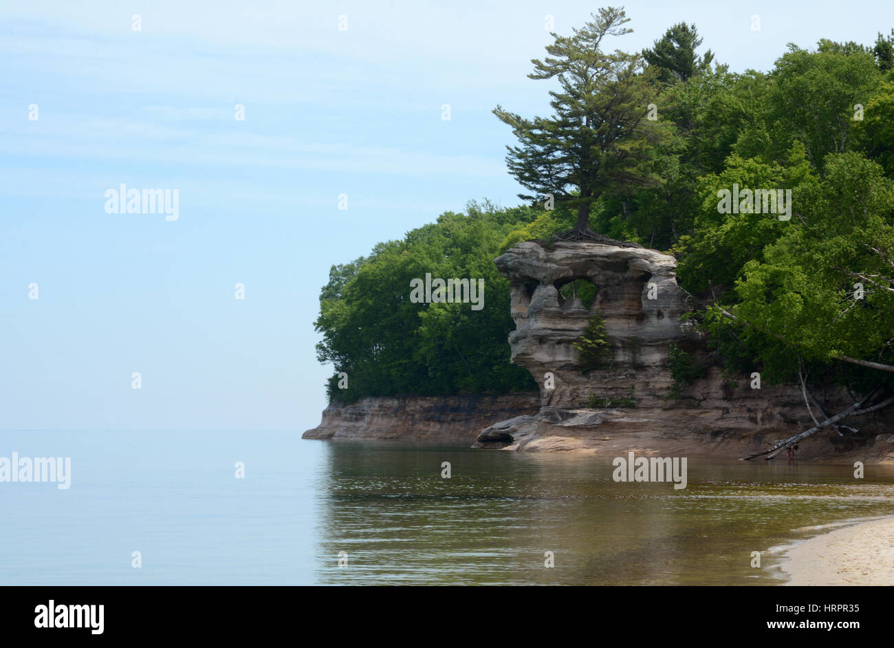 Il blu cielo incontra acqua calma sulla sinistra dell'immagine, e sulla destra è una formazione rocciosa con un'apertura ed è circondato da alberi. Foto Stock