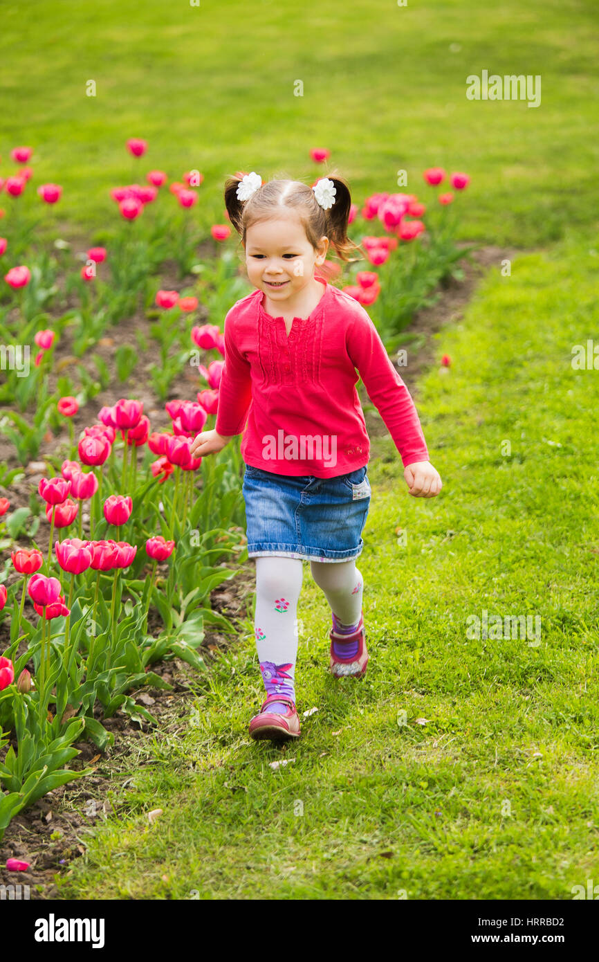 Carino divertente active bambina di quattro anni in esecuzione da soli allegramente fuori lungo aiuola con tulipani rosa in primavera soleggiata città park. Colore verticale p Foto Stock