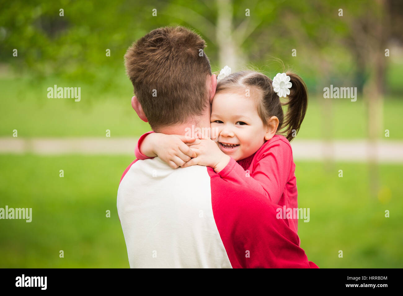 Closeup ritratto di famiglia felice. Poco funny girl di 4 anni di età che abbraccia il suo papà nella primavera del parco della città più verdi alberi ed erba sfondo. Horizo Foto Stock