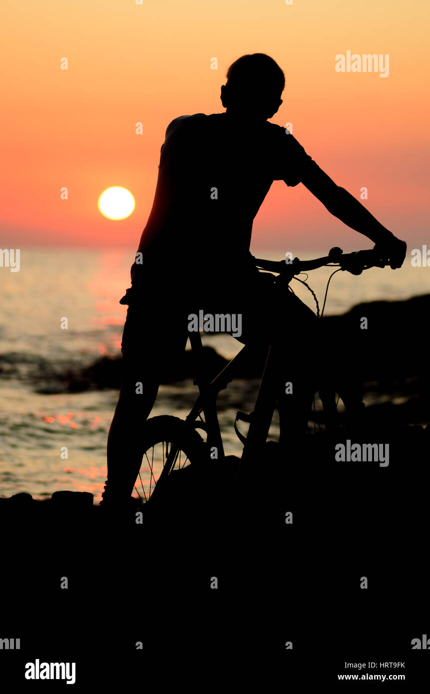 Silhouette di un giovane uomo in piedi con la bicicletta su una costa del mare e guardando il tramonto Foto Stock