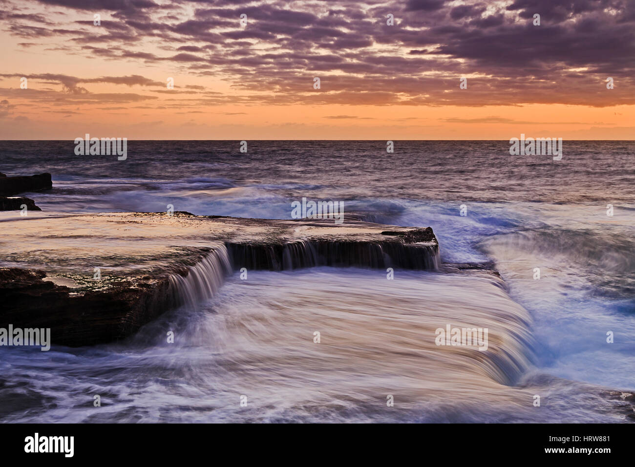 Colorato sunrise caldo da Maroubra Beach a Sydney. Navigare in flusso di onde piane su rocce di arenaria della costa australiana. Foto Stock
