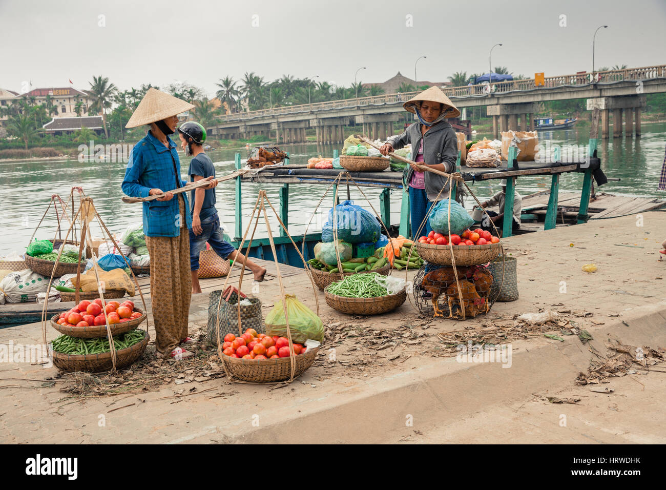 Hoi An, Vietnam - Marzo 14, 2014: Le donne nel cappello conico e l'uomo lo scarico di barca con merci per la vendita sul mercato dei prodotti freschi Foto Stock