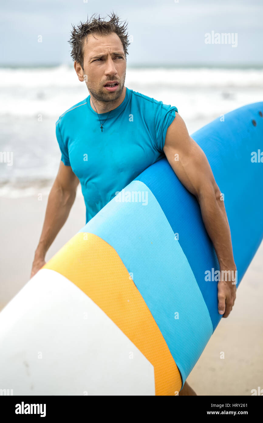 Bel ragazzo con i capelli disheveled e la stoppia sorge sulla spiaggia sulla sfondo sfocato di le onde del mare e del cielo. Egli indossa una ciano T-s Foto Stock