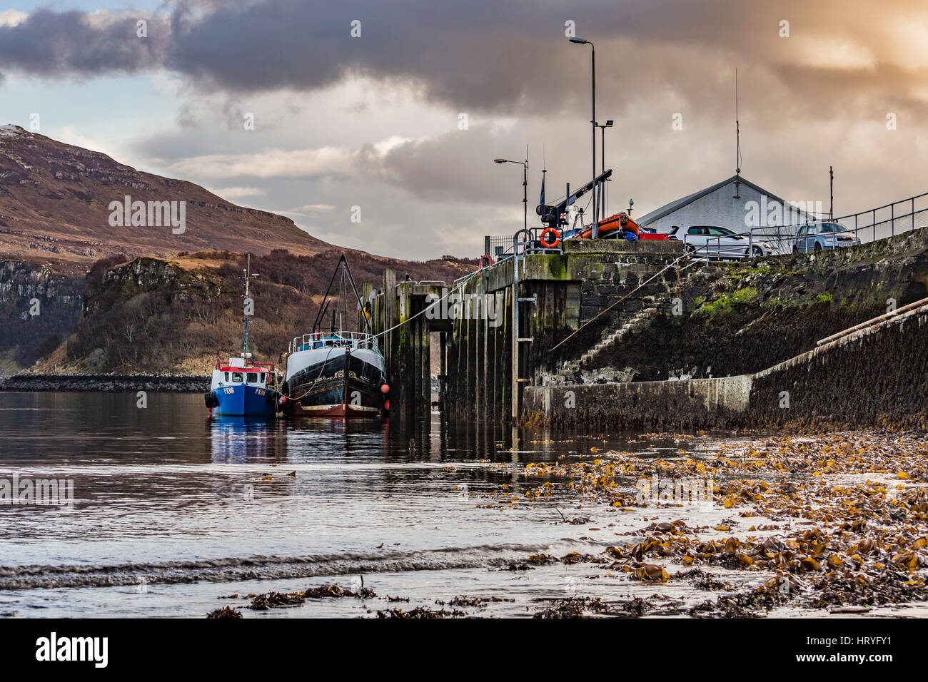 Ben Tianavaig attraverso Portree Harbour, sull'Isola di Skye in Scozia, contro un cielo drammatico, dietro alla fine del molo del porto e le barche Foto Stock