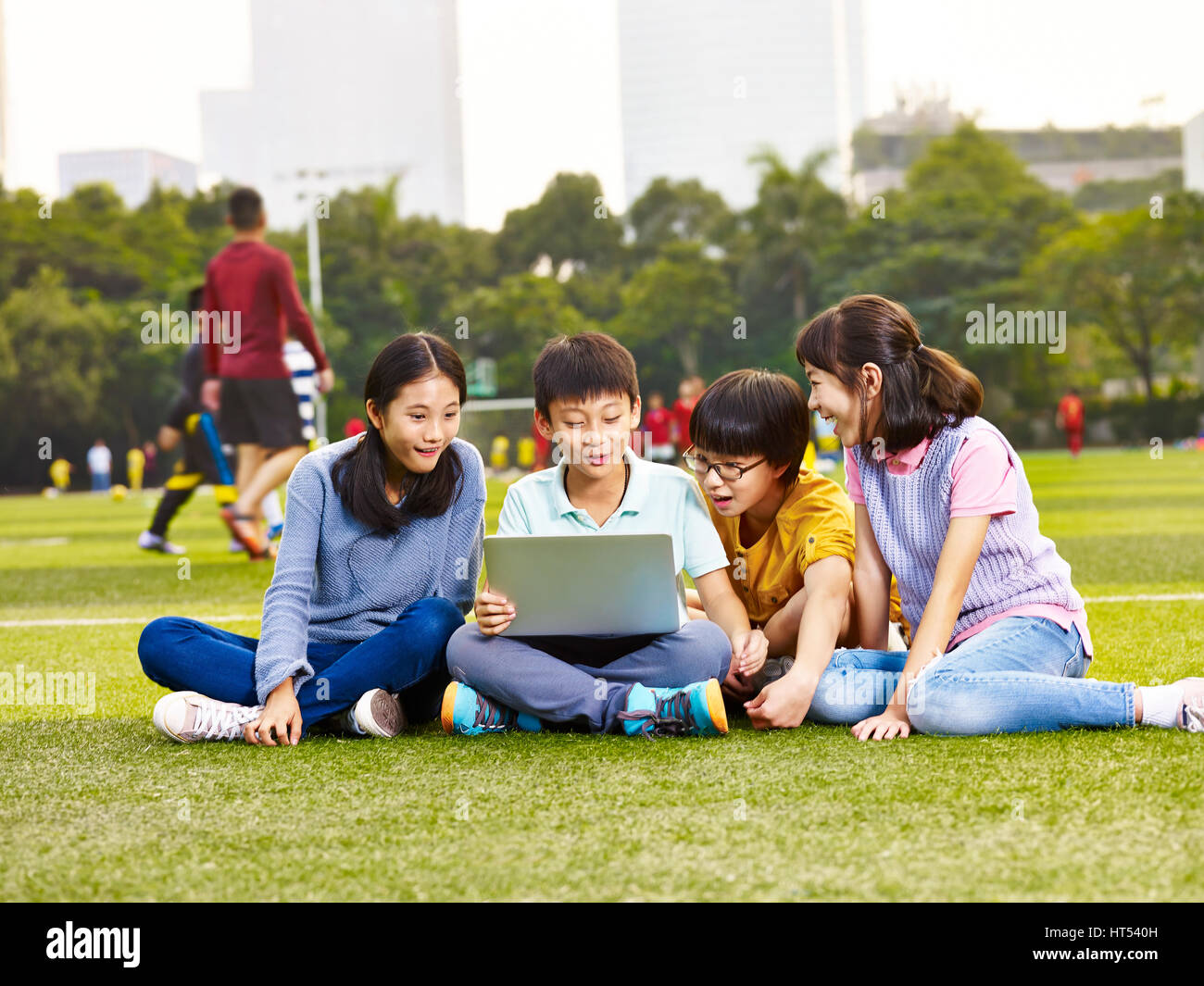 Gruppo di asiatici scuola elementare bambini seduti sul parco giochi erba guardando il computer portatile Foto Stock