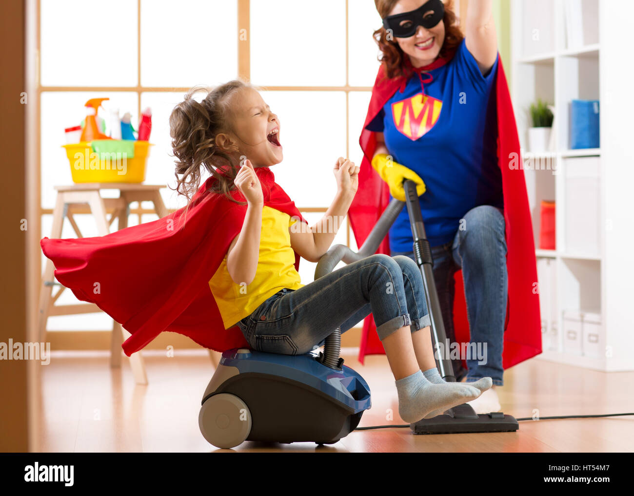 Il bambino e la madre vestita come supereroi utilizzando aspirapolvere in camera. Famiglia di mezza età della donna e bambino figlia hanno un divertimento durante la pulizia del pavimento. Foto Stock