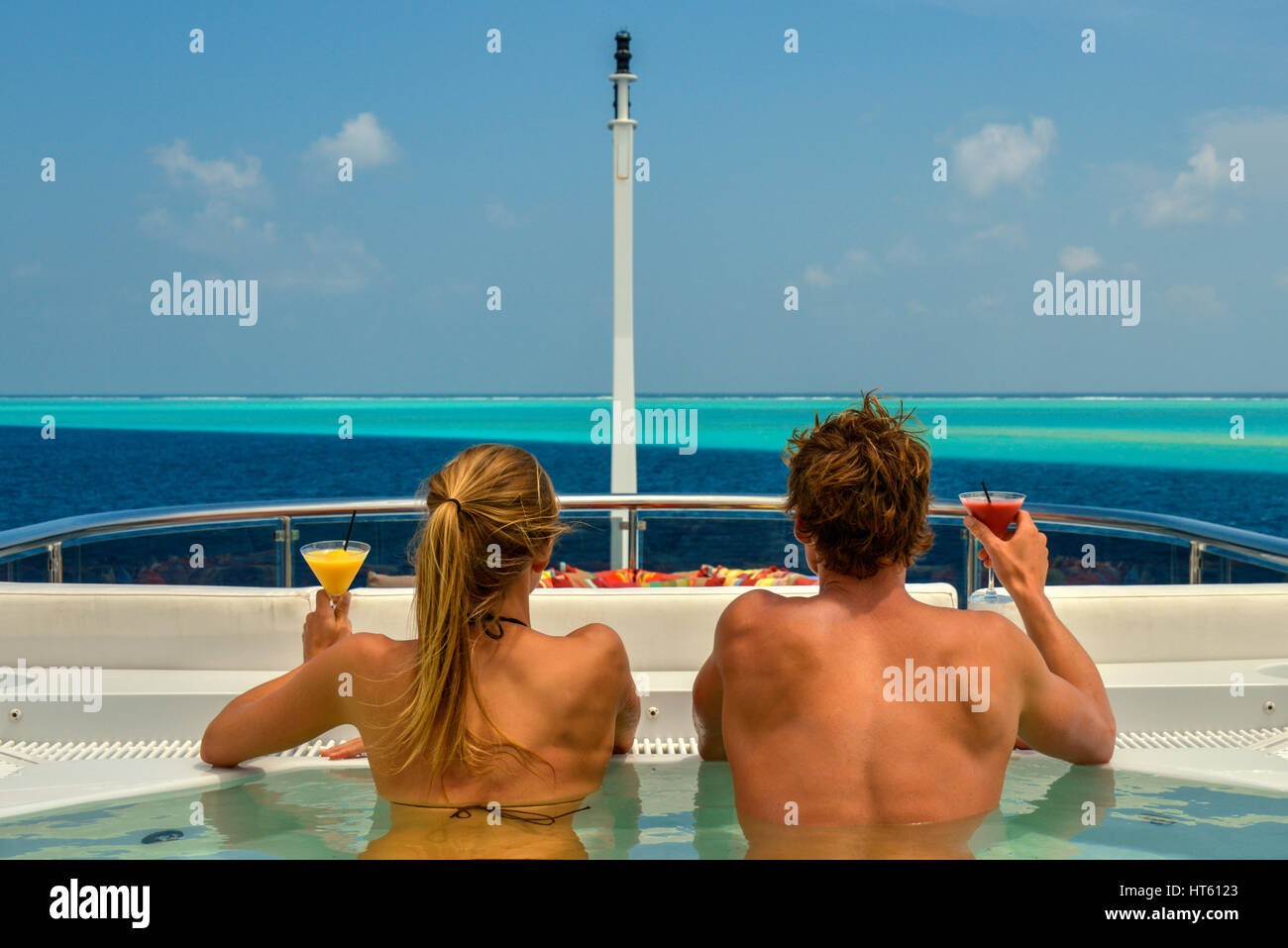 Una vista posteriore di una coppia bronzata e prendisole, con cocktail in mano, su uno yacht di lusso, che si affaccia su una barriera corallina verde blu Foto Stock