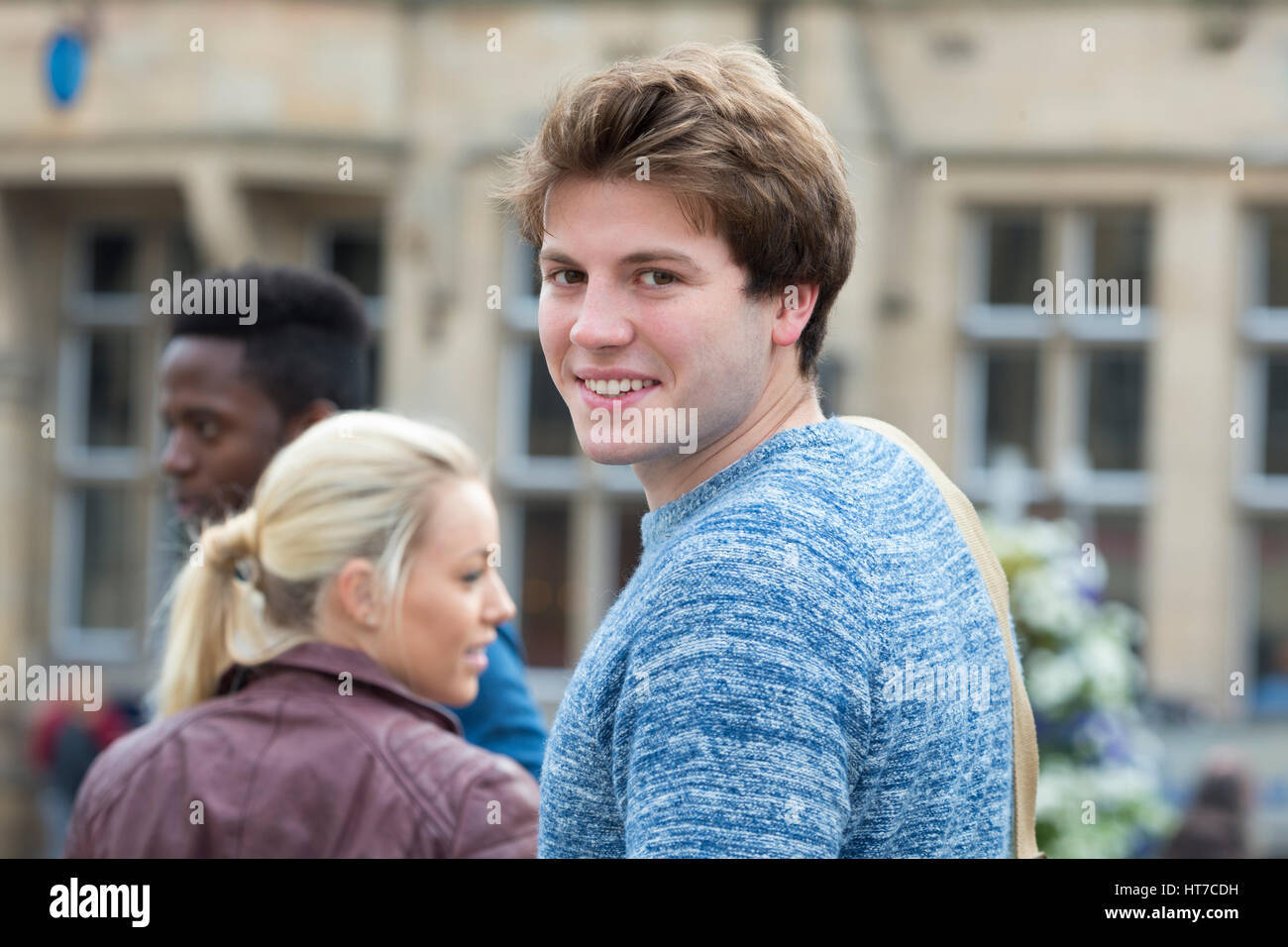 Un felice studente spire di sorridere alla telecamera come fa il suo modo per le sue lezioni. Egli sta portando il suo sacco sulle spalle e i suoi amici sono in b Foto Stock