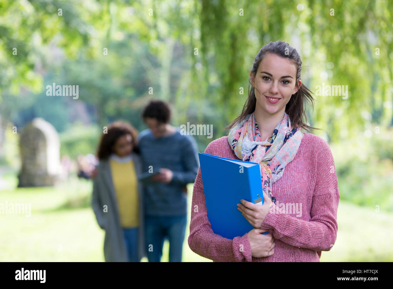 Un felice giovane donna sorride alla telecamera come essa si erge al di fuori tenendo i suoi libri. Un giovane è al di fuori della messa a fuoco in background utilizzando una tavoletta digitale. Foto Stock