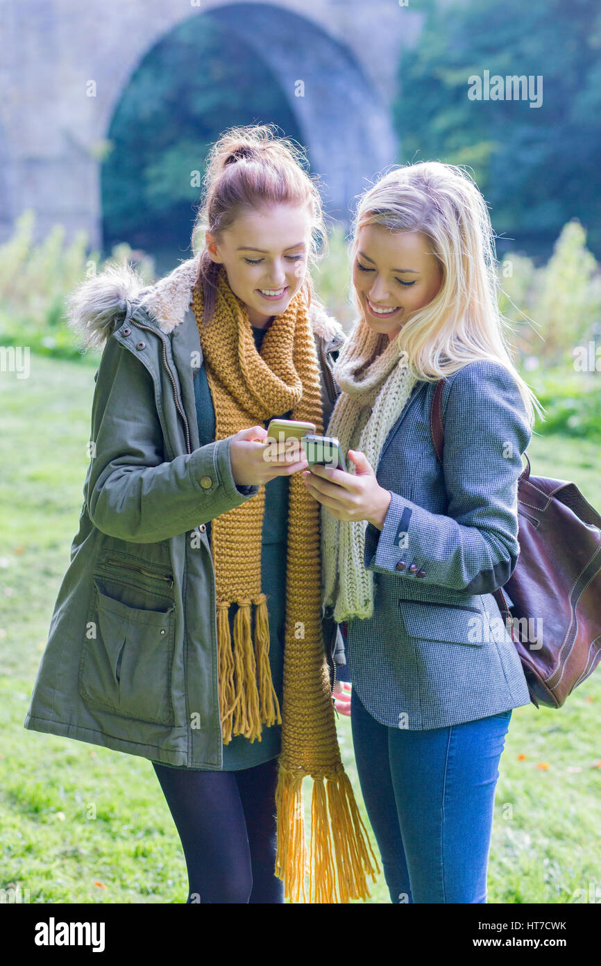 Due lieti studentesse sorriso come usano i loro telefoni cellulari insieme all'aperto. Essi sono avvolti fino a caldo in piedi vicino a un fiume. Foto Stock