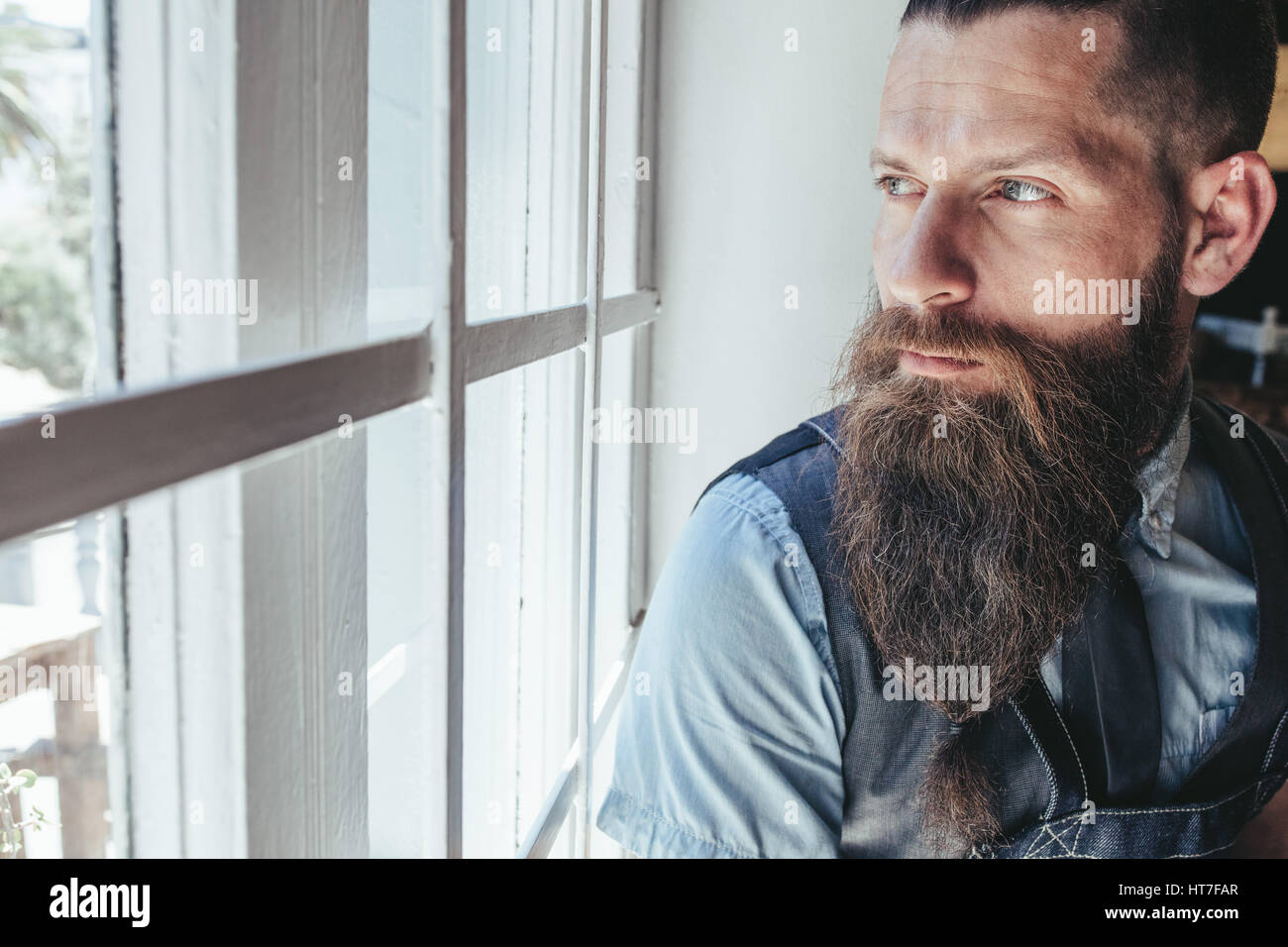 Close up di grave barbiere maschio con la barba lunga di guardare fuori dalla finestra e pensare. Foto Stock
