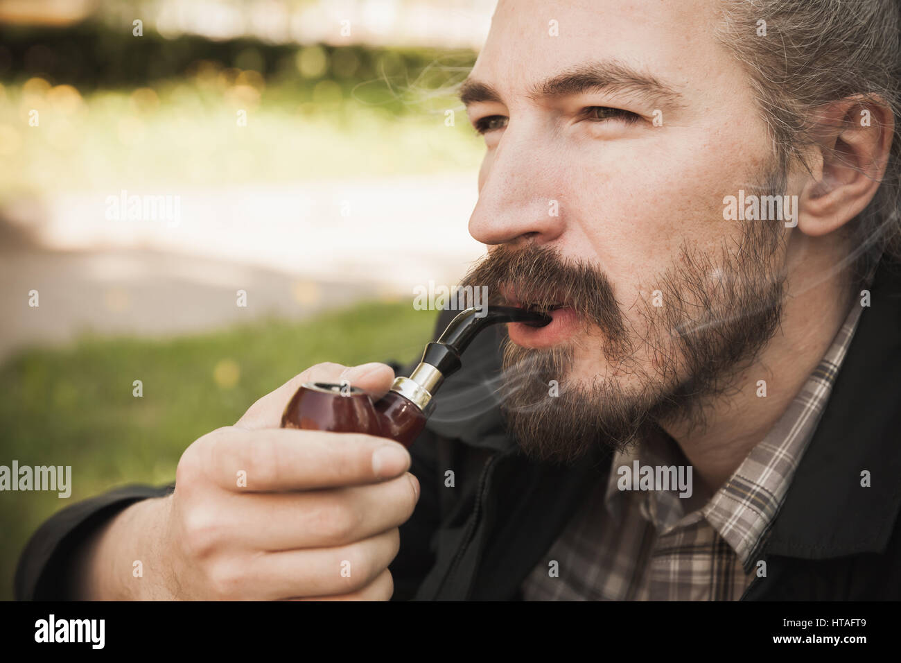Giovani asiatici grave uomo barbuto tubazione di fumare in estate park, vicino su ritratto Foto Stock