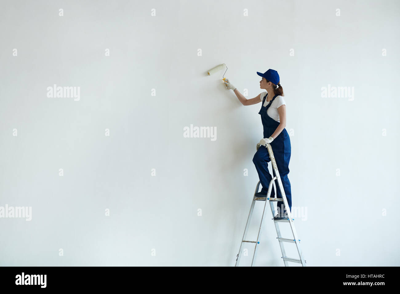 Casa-pittore in uniforme stando in piedi sulla scala e muro dipinto con il rullo Foto Stock
