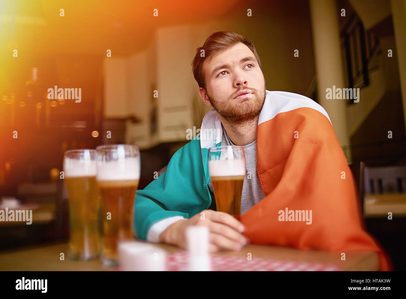 Ritratto di uomo barbuto avvolto in irlandese banner che guarda lontano premurosamente, seduti a tavola in pub con calici di birra artigianale Foto Stock