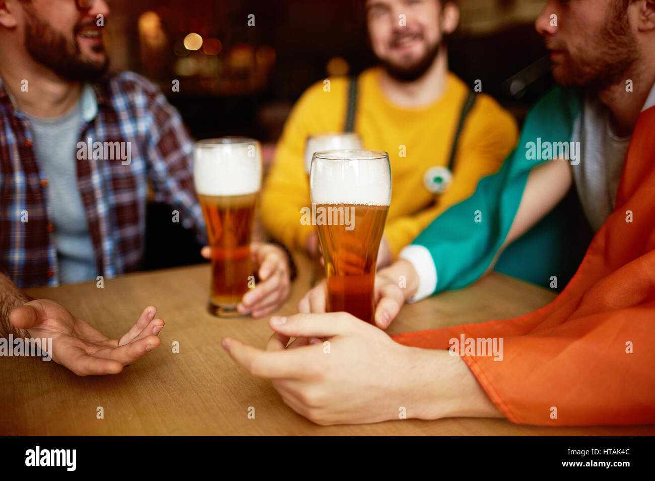 Closeup shot di alti bicchieri di birra chiara con giunto viscoso schiuma alta nelle mani di tre uomini seduti a tavola rotonda godendo cordiale incontro nel Pub Irlandese Foto Stock