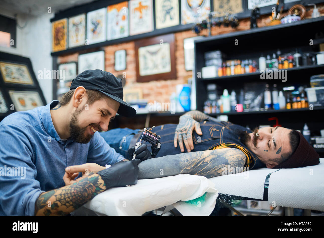 Felice tattooer giovani in ball-cap a lavorare con il cliente Foto Stock
