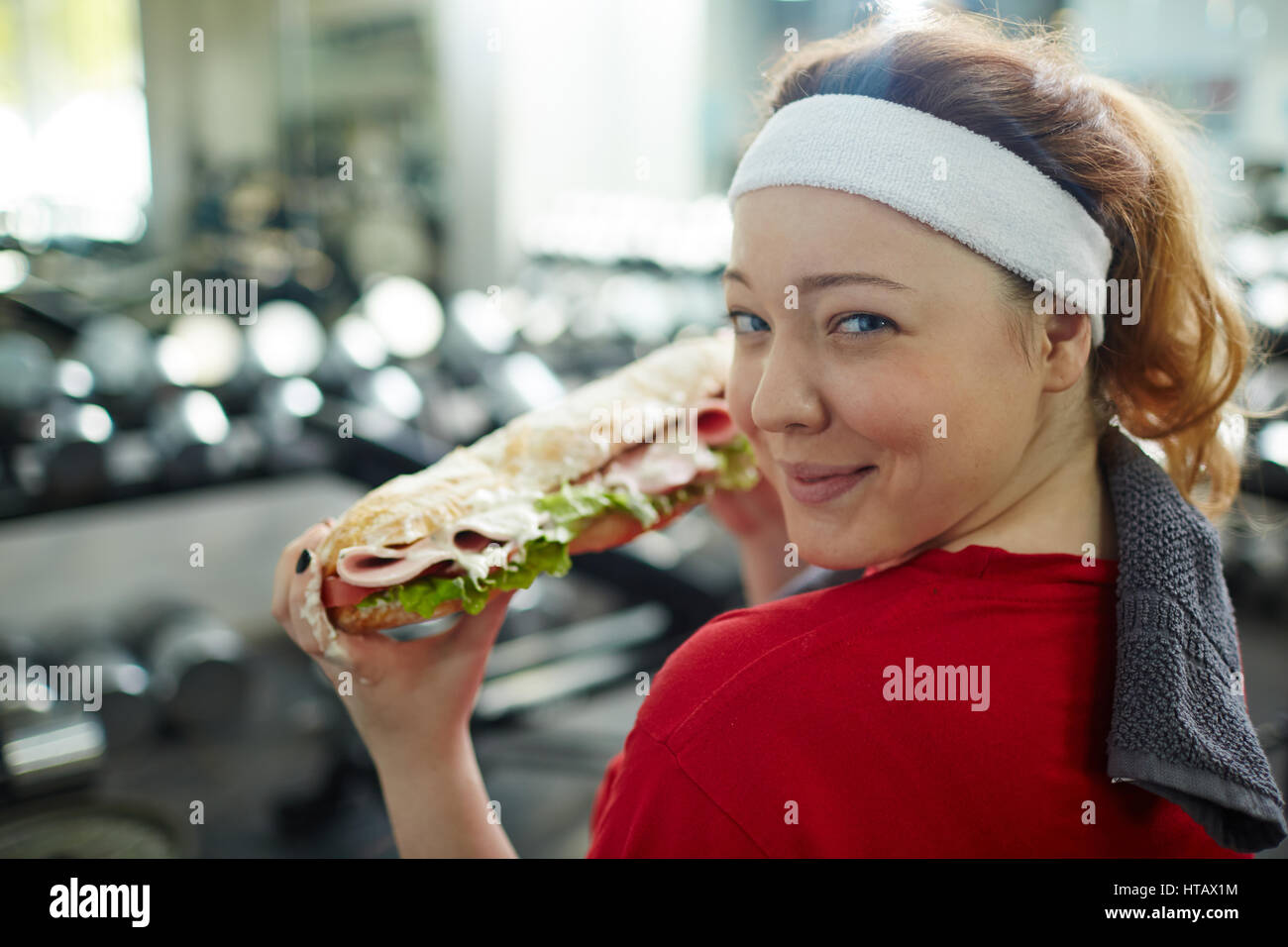 Ritratto di carino donna sovrappeso guardando sornione in telecamera, sorridente e tenuta grasso enorme panino mentre si lavora in palestra, lotta di perdita di peso quando Foto Stock