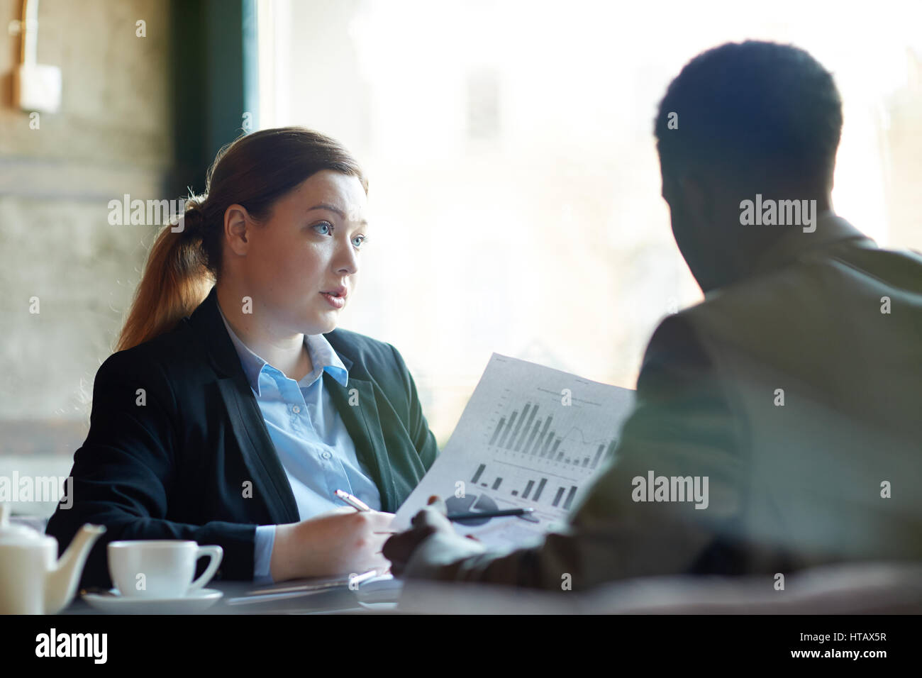 Due uomini di affari che durante la riunione di lavoro in moderno Cafe: giovane donna professionale per spiegare i dati statistici tenendo i documenti con i grafici a parte maschio Foto Stock