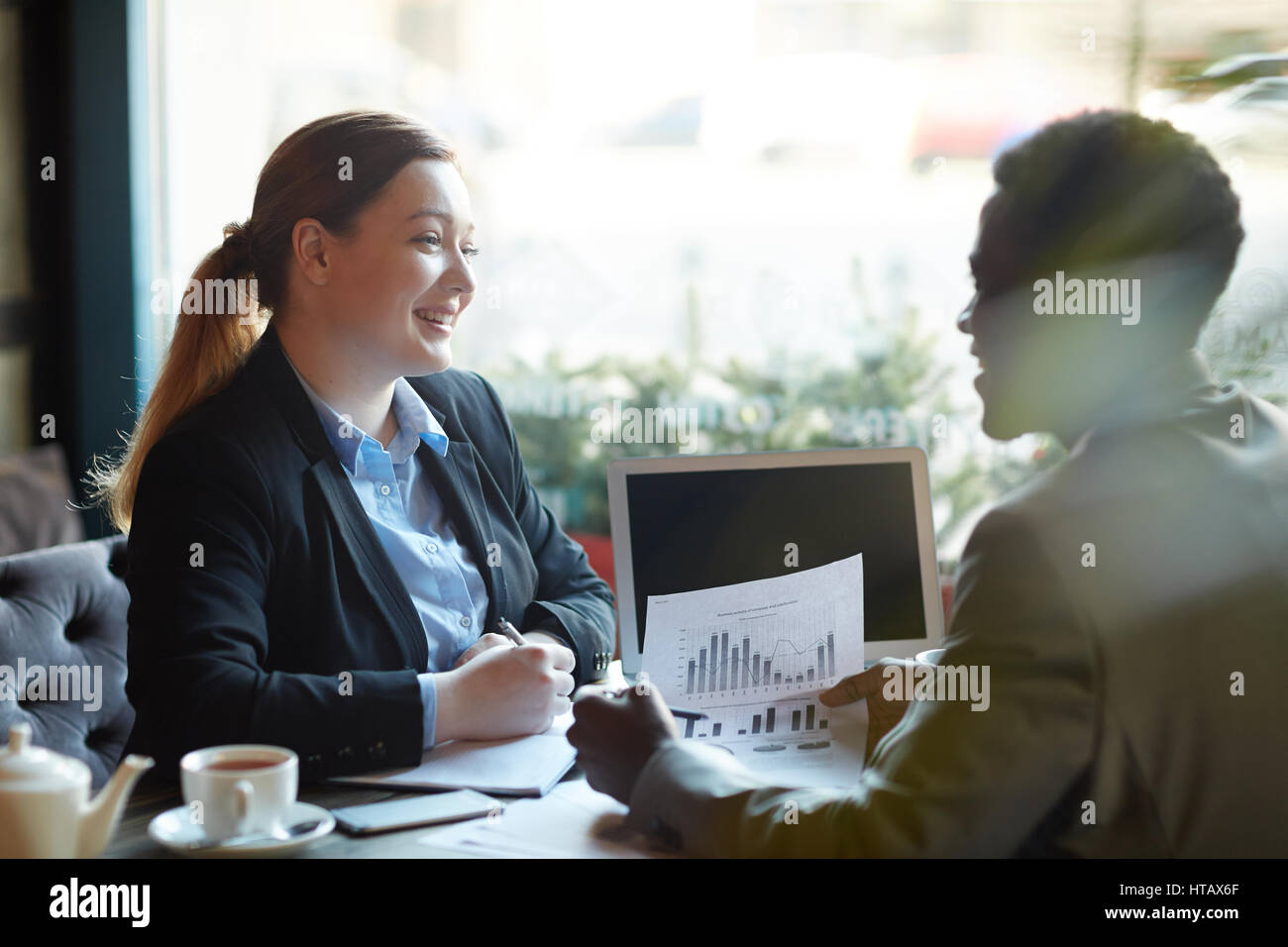 Due uomini di affari che durante la riunione di lavoro in moderno Cafe: giovane professionista donna sorridente in paesi africani partner americano mentre si lavora con le statistiche doc Foto Stock