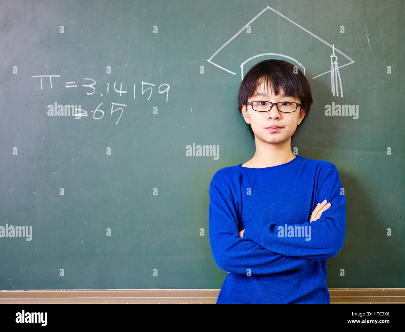 Asian scuola elementare ragazzo con gli occhiali in piedi sotto un cappello di dottorato disegnato con un gessetto sulla lavagna. Foto Stock