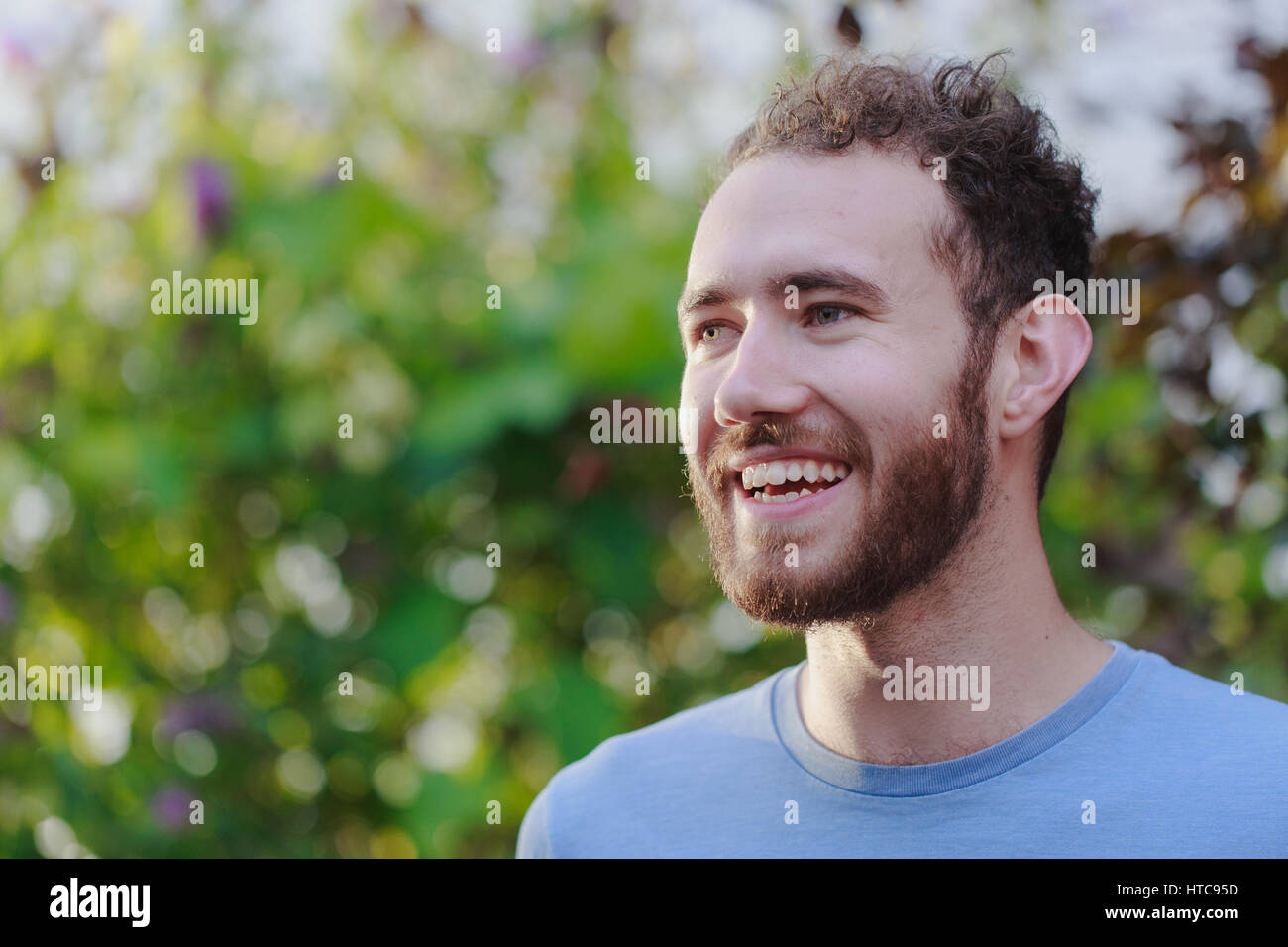 Sorridente giovane con parentesi marrone capelli e barba, hipster, indossa una maglietta blu, ritratto in giardino. Golden ore di luce Foto Stock