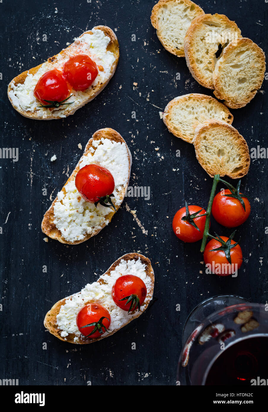 Toast con formaggio bianco diffuso e arrosto di pomodori ciliegia e bicchiere di vino rosso su sfondo nero. Vista superiore Foto Stock