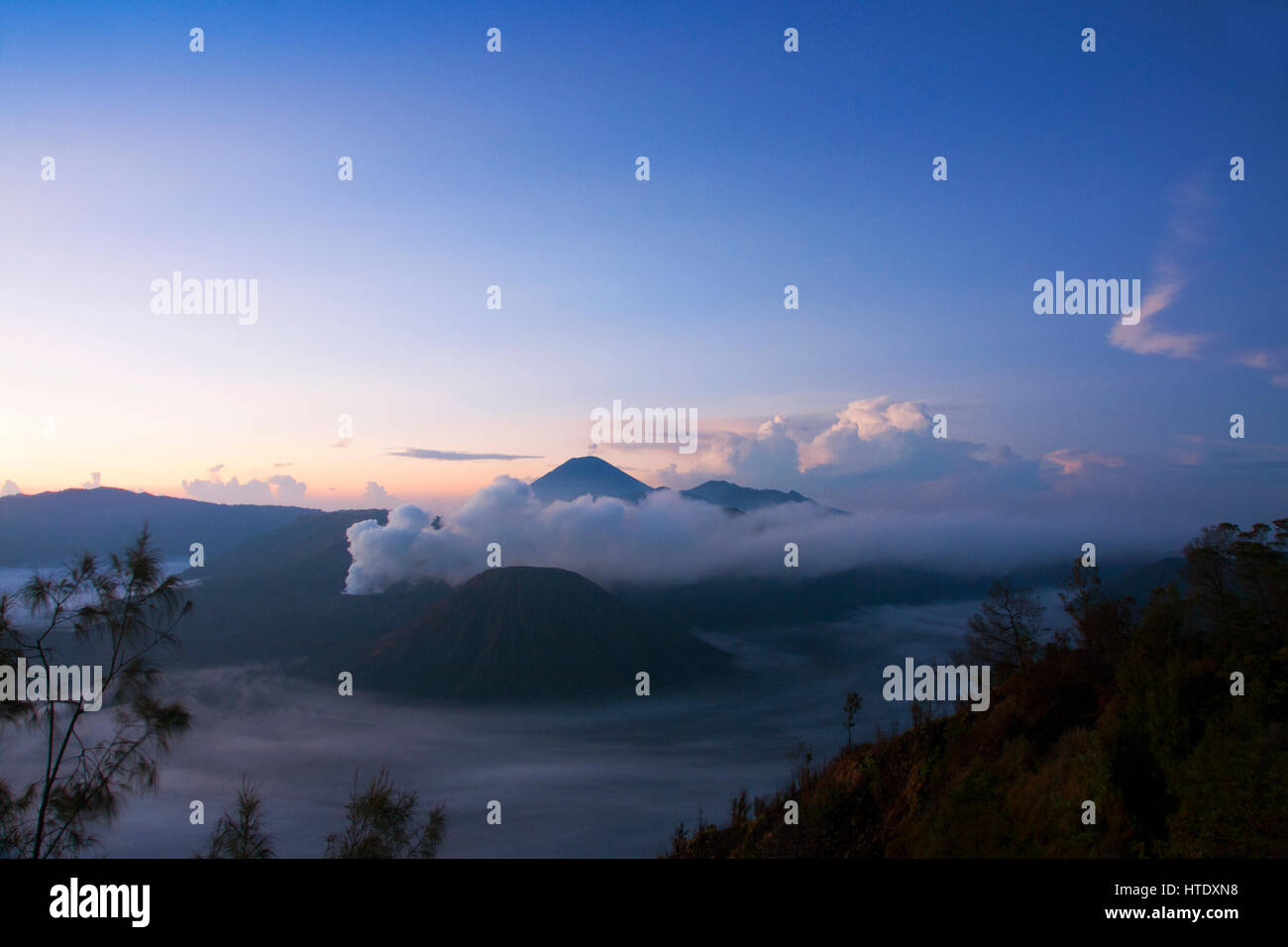 Fumo bianco in uscita dei vulcani circondato da nuvole bianche di nebbia e un cielo azzurro visto ad una distanza nel pomeriggio da Mount Penanjakan Foto Stock