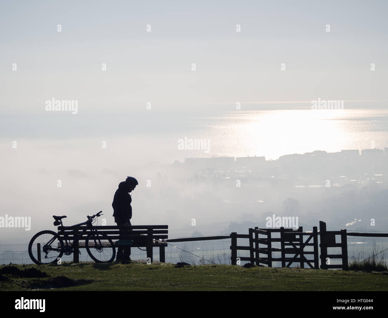 profilo di una silhouette di un elmetto da ciclista solitario in piedi da solo sulla parte superiore Di Downs collina con stile guardando verso la nebbia nebbia nella valle e mare sottostante Foto Stock