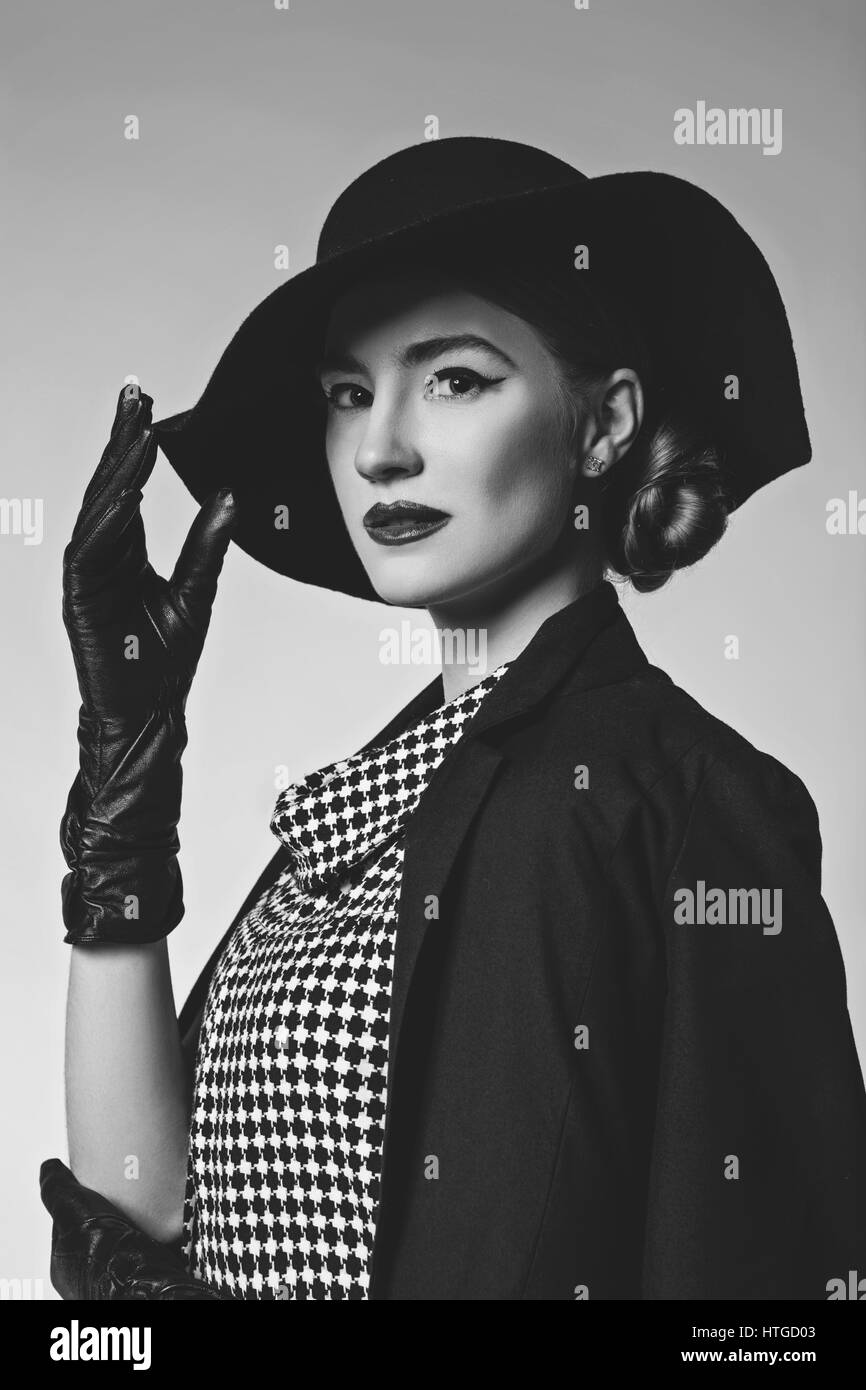 Bella giovane donna con labbra rosse per il make-up retrò in bianco e nero vestito, cappello, giacca e guanti in pelle in piedi su sfondo grigio. Monocromatico. Foto Stock