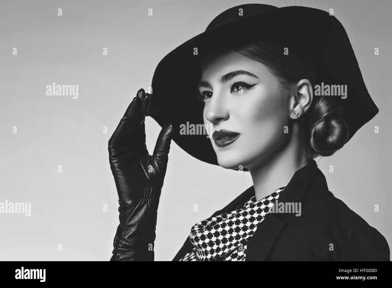 Bella giovane donna con labbra rosse per il make-up retrò in bianco e nero vestito, cappello, giacca e guanti in pelle in piedi su sfondo grigio. Monocromatico. Co Foto Stock